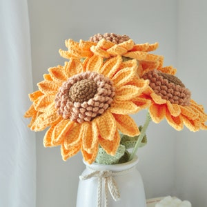 May include: A bouquet of handmade crochet sunflowers in a white vase. The sunflowers have bright yellow-orange petals and brown centers. The vase is white with a tied bow. The flowers are a decorative item.