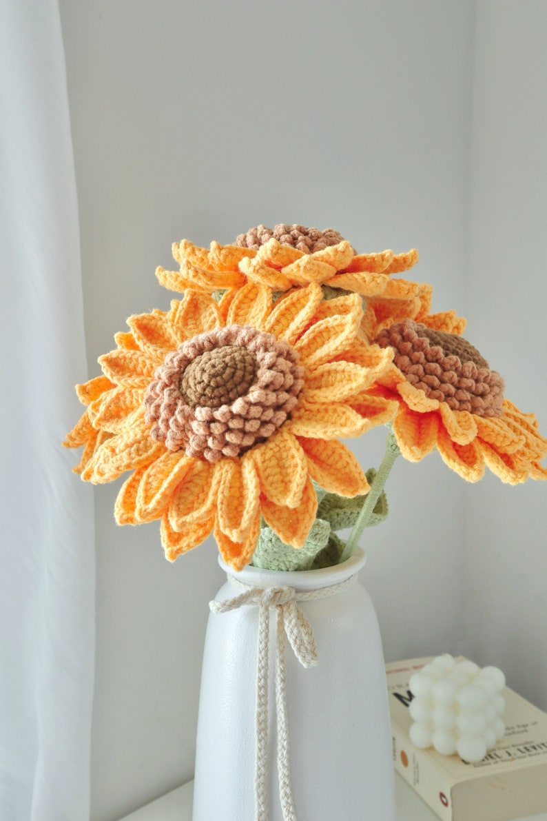 May include: A bouquet of handmade crochet sunflowers in a white vase. The sunflowers have bright yellow-orange petals and brown centers. The vase is white with a tied bow. The flowers are a decorative item.