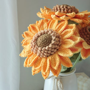 May include: A bouquet of handmade crochet sunflowers in a white vase. The sunflowers have bright yellow petals and brown centers. The vase is white with a decorative rope tie. The flowers are a cheerful home decor item.
