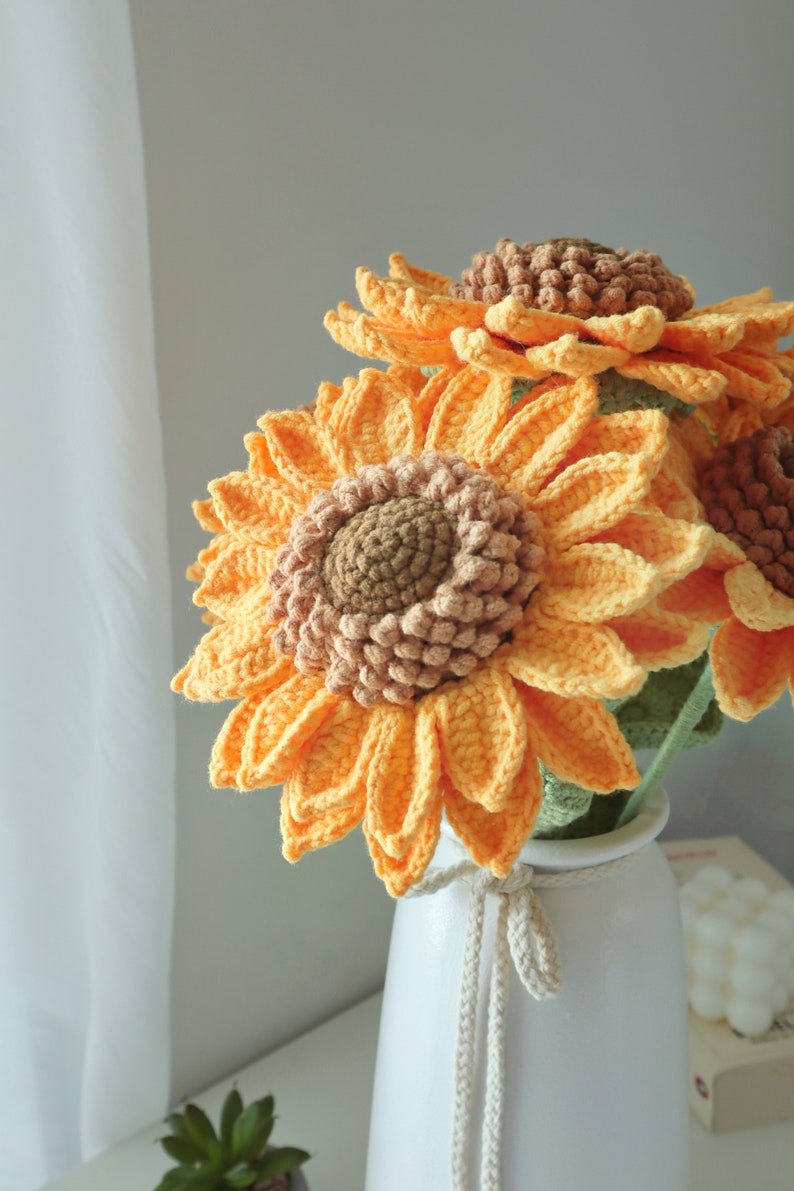 May include: A bouquet of handmade crochet sunflowers in a white vase. The sunflowers have bright yellow petals and brown centers. The vase is white with a decorative rope tie. The flowers are a cheerful home decor item.