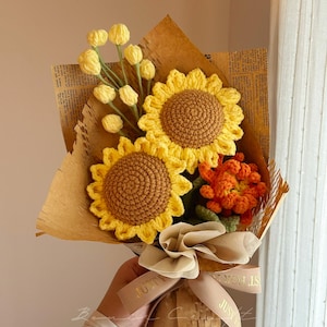 Puede incluir: Un ramo de flores de ganchillo con dos grandes girasoles amarillos de centro marr&oacute;n, un racimo de peque&ntilde;os capullos amarillos sobre tallos verdes y una flor naranja texturizada, envuelto en papel marr&oacute;n con una cinta beige con el texto JUST FOR YOU.