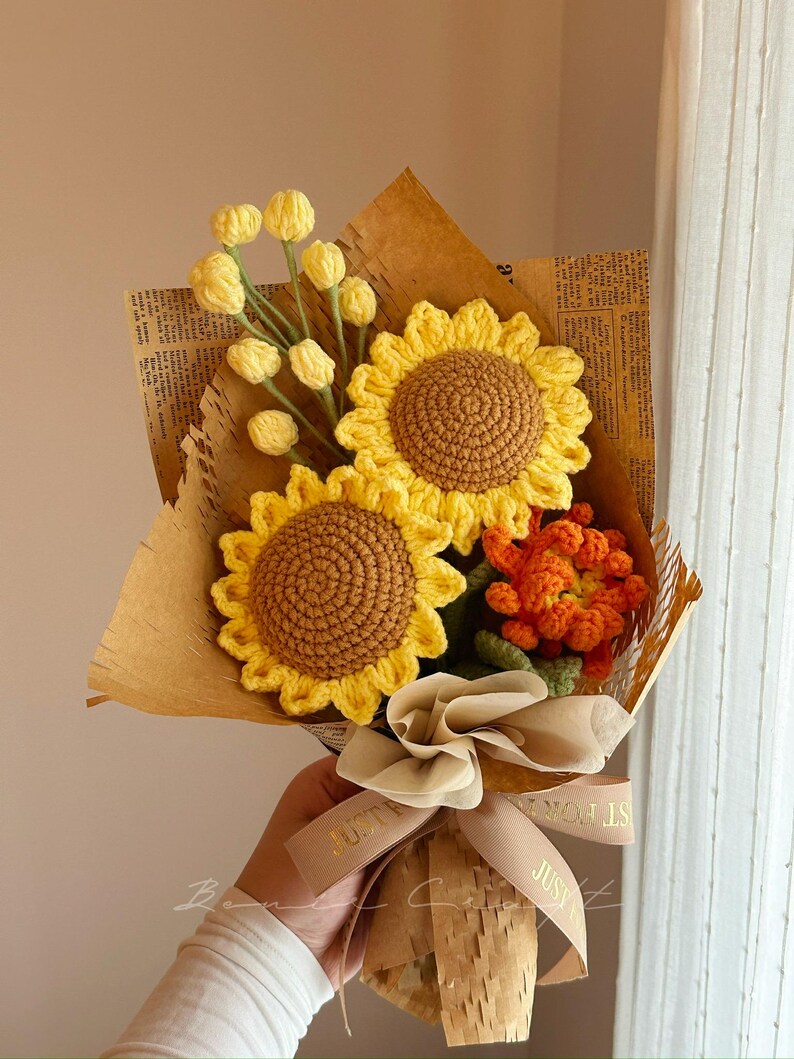 Puede incluir: Un ramo de flores de ganchillo con dos grandes girasoles amarillos de centro marr&oacute;n, un racimo de peque&ntilde;os capullos amarillos sobre tallos verdes y una flor naranja texturizada, envuelto en papel marr&oacute;n con una cinta beige con el texto JUST FOR YOU.