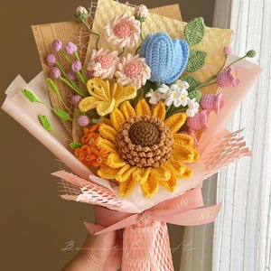 Puede incluir: Un ramo de flores de ganchillo con un gran girasol amarillo, un tulip&aacute;n azul, flores rosas y hojas verdes. El arreglo est&aacute; envuelto en papel rosa y malla decorativa, atado con una cinta rosa con el texto impreso FOR YOU.