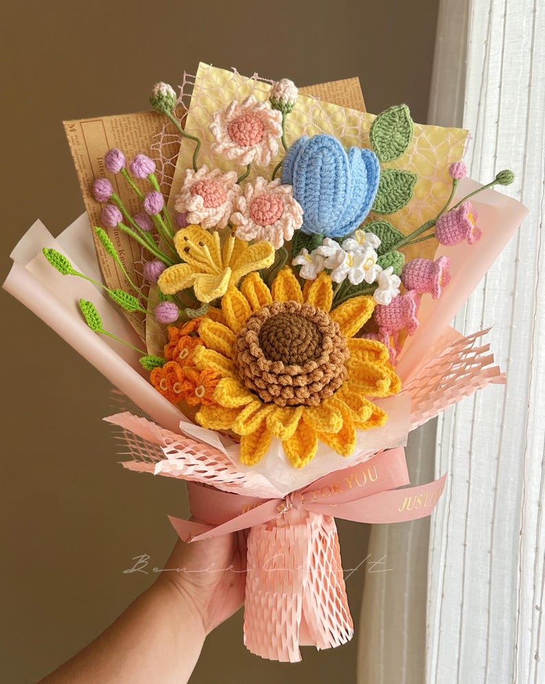 Puede incluir: Un ramo de flores de ganchillo con un gran girasol amarillo, un tulip&aacute;n azul, flores rosas y hojas verdes. El arreglo est&aacute; envuelto en papel rosa y malla decorativa, atado con una cinta rosa con el texto impreso FOR YOU.