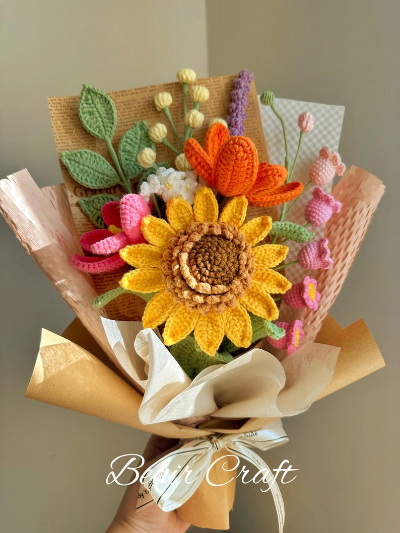 May include: A handcrafted bouquet of crochet flowers, including a large yellow sunflower, orange lotus, and pink blossoms. The bouquet is wrapped in brown paper and tied with a white ribbon. The text "Beir Craft" is visible on the wrapping.