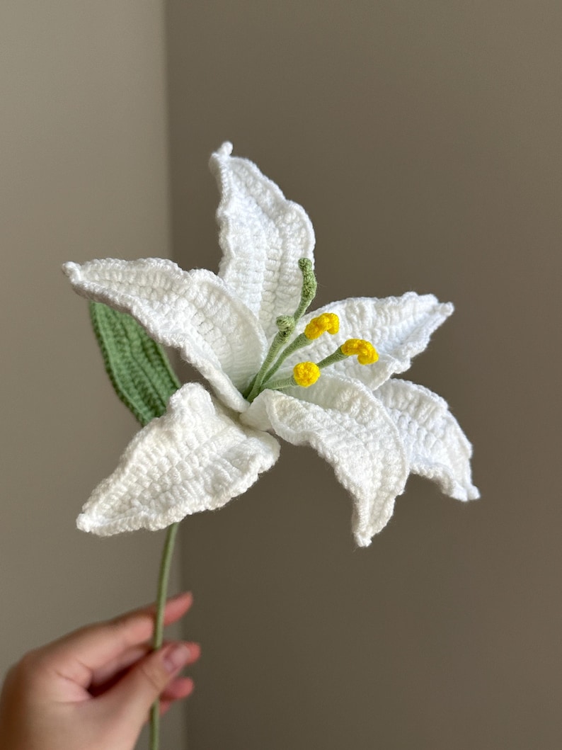 Puede incluir: Una flor de lirio tejida a mano en crochet con seis p&eacute;talos blancos de textura marcada. El centro presenta estambres de color verde claro rematados con puntas de polen amarillo brillante. Una hoja verde tejida est&aacute; unida al tallo.