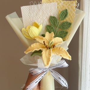 Puede incluir: Un ramo de flores de ganchillo que incluye un lirio amarillo y un tulip&aacute;n amarillo, acompa&ntilde;ados de hojas verdes tejidas. El ramo est&aacute; envuelto en papel blanco y amarillo con una cinta blanca impresa con el texto Always By Your Side.