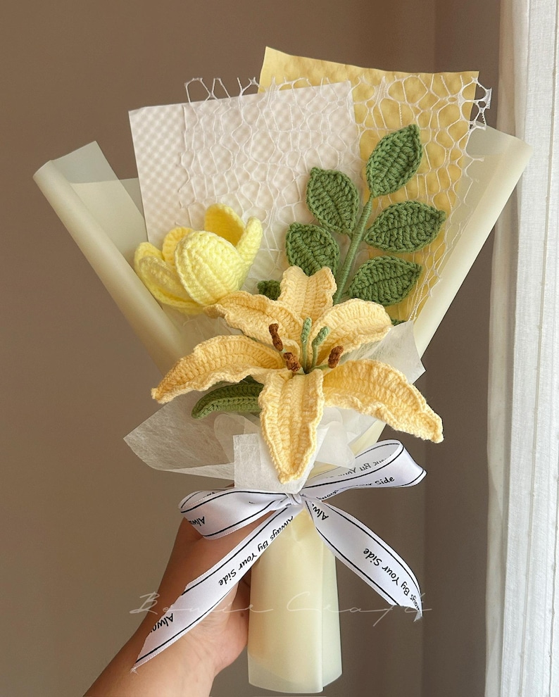 Puede incluir: Un ramo de flores de ganchillo que incluye un lirio amarillo y un tulip&aacute;n amarillo, acompa&ntilde;ados de hojas verdes tejidas. El ramo est&aacute; envuelto en papel blanco y amarillo con una cinta blanca impresa con el texto Always By Your Side.