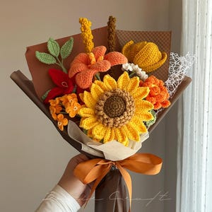 Puede incluir: Un ramo de flores de ganchillo que presenta un gran girasol amarillo, lirios naranjas y varias flores peque&ntilde;as texturizadas en tonos rojos, amarillos y verdes. El arreglo est&aacute; envuelto en papel marr&oacute;n con una cinta naranja decorativa en la base.