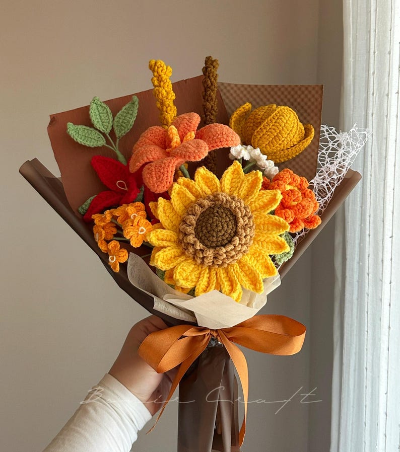 Puede incluir: Un ramo de flores de ganchillo que presenta un gran girasol amarillo, lirios naranjas y varias flores peque&ntilde;as texturizadas en tonos rojos, amarillos y verdes. El arreglo est&aacute; envuelto en papel marr&oacute;n con una cinta naranja decorativa en la base.