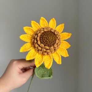 May include: A handmade crochet sunflower with bright yellow petals, a brown center, and a green stem and leaf. The sunflower is held against a gray background, showcasing the detailed craftsmanship of the floral design.