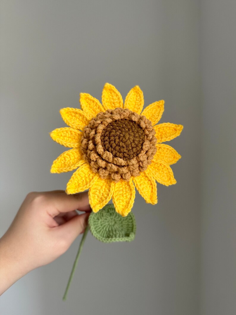 May include: A handmade crochet sunflower with bright yellow petals, a brown center, and a green stem and leaf. The sunflower is held against a gray background, showcasing the detailed craftsmanship of the floral design.