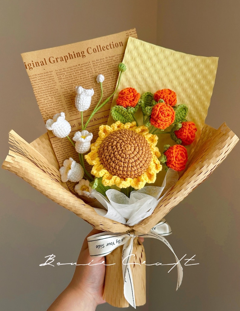 Puede incluir: Un ramo de flores de ganchillo con un gran girasol amarillo, varias flores blancas peque&ntilde;as en forma de campana y flores naranjas similares a rosas con hojas verdes. El arreglo est&aacute; envuelto en papel marr&oacute;n y una hoja con texto impreso.