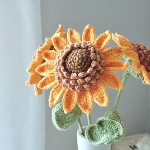 May include: A bouquet of handmade crochet sunflowers in a white vase. The sunflowers have orange petals, brown centers, and green leaves. The vase is tied with twine. A decorative item for the home.