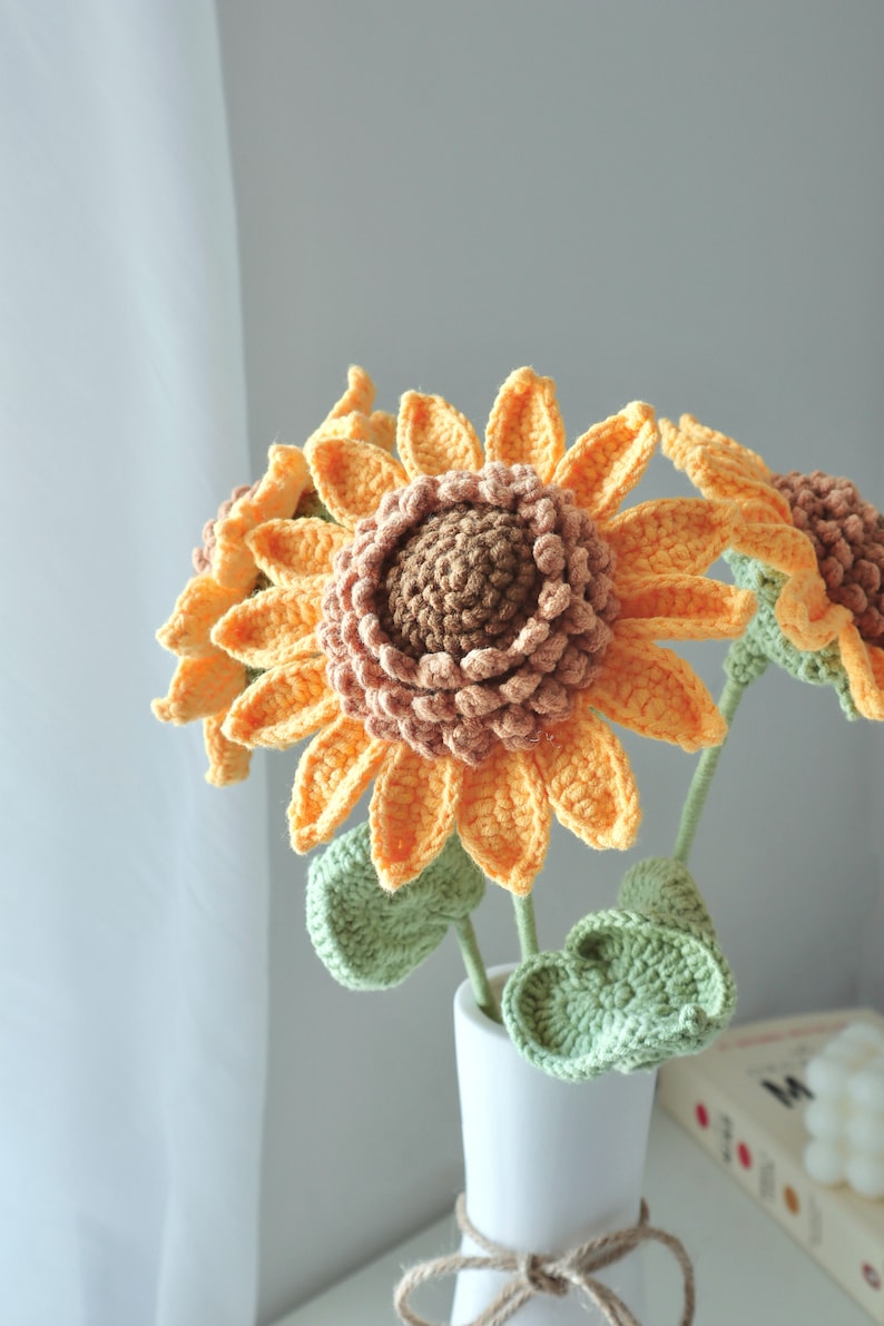May include: A bouquet of handmade crochet sunflowers in a white vase. The sunflowers have orange petals, brown centers, and green leaves. The vase is tied with twine. A decorative item for the home.