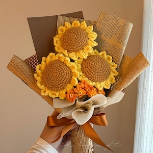 Puede incluir: Un ramo de tres girasoles de ganchillo con p&eacute;talos amarillos y centros marrones, acompa&ntilde;ados de peque&ntilde;as flores de ganchillo naranjas. El arreglo est&aacute; envuelto en papel marr&oacute;n y papel de peri&oacute;dico, atado con una cinta de sat&eacute;n marr&oacute;n.