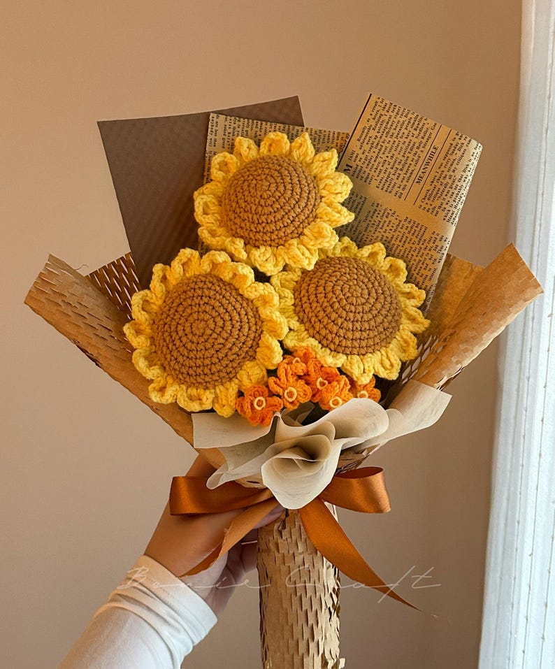 Puede incluir: Un ramo de tres girasoles de ganchillo con p&eacute;talos amarillos y centros marrones, acompa&ntilde;ados de peque&ntilde;as flores de ganchillo naranjas. El arreglo est&aacute; envuelto en papel marr&oacute;n y papel de peri&oacute;dico, atado con una cinta de sat&eacute;n marr&oacute;n.