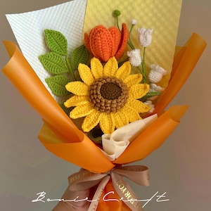 Puede incluir: Un ramo de flores de ganchillo que presenta un gran girasol amarillo con centro marr&oacute;n, un tulip&aacute;n naranja y peque&ntilde;as flores blancas en forma de campana con hojas verdes. El ramo est&aacute; envuelto en papel naranja y blanco con una cinta impresa.