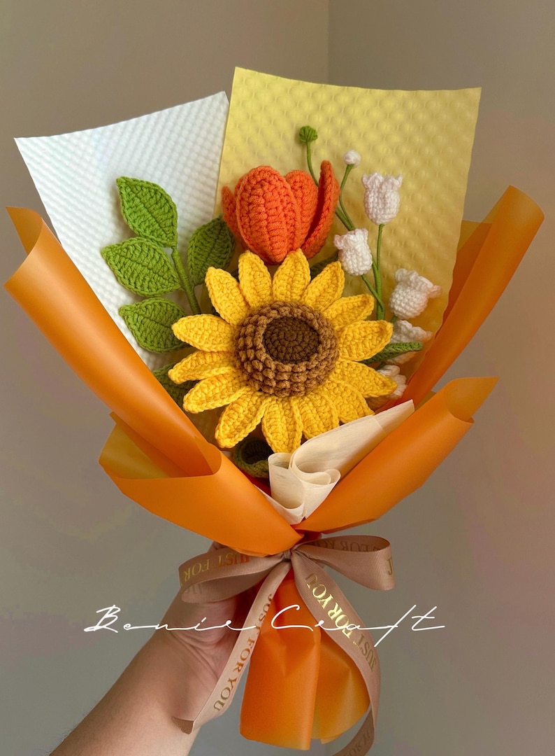 Puede incluir: Un ramo de flores de ganchillo que presenta un gran girasol amarillo con centro marr&oacute;n, un tulip&aacute;n naranja y peque&ntilde;as flores blancas en forma de campana con hojas verdes. El ramo est&aacute; envuelto en papel naranja y blanco con una cinta impresa.