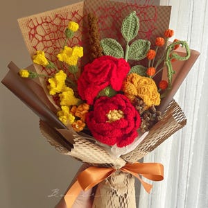 Puede incluir: Un ramo de flores de ganchillo hecho a mano con rosas rojas vibrantes, flores amarillas y bayas naranjas. El arreglo incluye hojas y tallos verdes tejidos, envuelto en papel marr&oacute;n con un lazo naranja decorativo en la base.