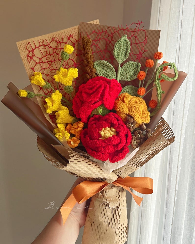 Puede incluir: Un ramo de flores de ganchillo hecho a mano con rosas rojas vibrantes, flores amarillas y bayas naranjas. El arreglo incluye hojas y tallos verdes tejidos, envuelto en papel marr&oacute;n con un lazo naranja decorativo en la base.