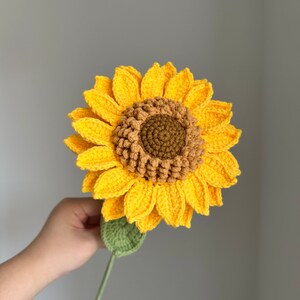 May include: A handmade crochet sunflower with bright yellow petals, a brown center, and a green stem and leaf. The flower is held up against a gray background.