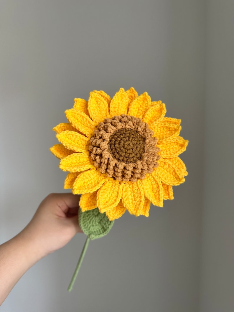 May include: A handmade crochet sunflower with bright yellow petals, a brown center, and a green stem and leaf. The flower is held up against a gray background.