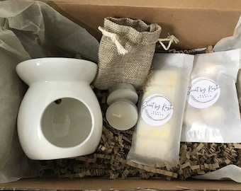May include: A white ceramic wax warmer with two white tea lights, two small bags of wax melts labelled "Scent by Kestie Baby Powder", and a small hessian bag. The items are in a brown cardboard box with brown shredded paper.