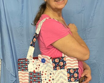 May include: A patchwork tote bag with a patriotic theme. The bag features squares of red, white, and blue fabric with stars, stripes, and the text "U.S.A.". The bag has a blue button closure and a white strap with blue and red accents.