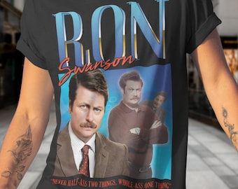 May include: A black t-shirt with a vintage-style graphic of Ron Swanson from the TV show Parks and Recreation. The graphic features a blue and gold text overlay that reads "Ron Swanson" and a quote from the show: "Never half-ass two things, whole-ass one thing."