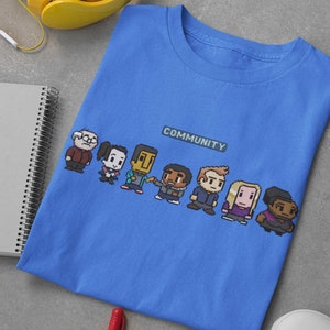 Könnte beinhalten: Ein blaues T-Shirt mit einer pixeligen Grafik der Figuren aus der Fernsehserie "Community" und dem Wort "COMMUNITY" auf der Vorderseite.