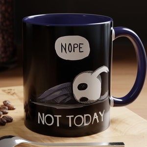 May include: A black and blue ceramic mug with a white graphic of a character from the video game Hollow Knight. The mug has the text "NOPE" in a speech bubble and "NOT TODAY" below the character.