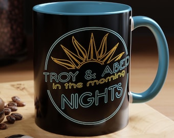 May include: Black ceramic mug with a blue rim and handle. The mug features a yellow sun graphic with the text "Troy & Abed in the morning nights" in a neon blue outline.