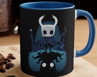 May include: A blue and black ceramic coffee mug with a graphic of a white knight character from the video game Hollow Knight. The knight is standing in front of a dark, shadowy figure.