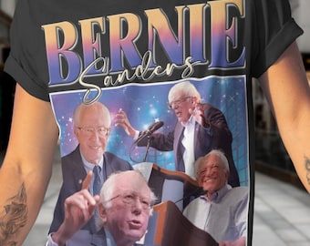 Puede incluir: Una camiseta negra con un estampado gráfico de Bernie Sanders. El gráfico presenta un collage de imágenes de Bernie Sanders en diferentes poses, incluyendo él hablando en un mitin y haciendo un pulgar hacia arriba. El texto "Bernie Sanders" está impreso en una fuente retro sobre el collage.