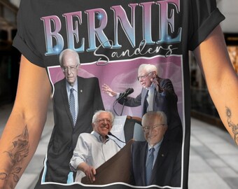May include: A black t-shirt with a vintage-style graphic of Bernie Sanders. The graphic features a collage of photos of Sanders, including one of him speaking at a podium. The text "Bernie Sanders" is printed in pink and blue letters above the graphic.