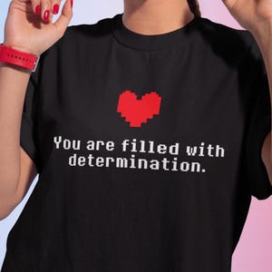 Könnte beinhalten: Schwarzes T-Shirt mit einem roten Pixelherz und dem Text "You are filled with determination."