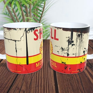 Puede incluir: Dos tazas de cerámica blanca con un diseño vintage de Shell Motor Oil. Las tazas presentan un esquema de color rojo, amarillo y blanco con un aspecto desgastado y oxidado. El texto "Shell Motor Oil" está impreso en las tazas, junto con las medidas "0.32 US Quart" y "0.3 Litre".