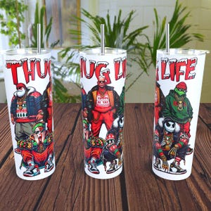 Puede incluir: Tres vasos blancos con un diseño de "Thug Life" con temática navideña. Los vasos presentan personajes de dibujos animados con gorros de Papá Noel y suéteres navideños. El texto "Thug Life" está impreso en cada vaso.