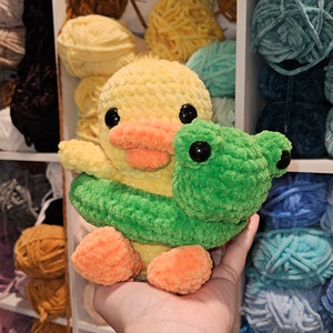 May include: A crocheted yellow duck wearing a green crocheted inner tube with a green crocheted frog on top. The duck and frog are holding onto the inner tube.