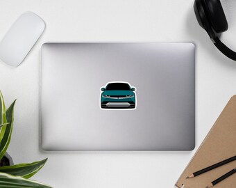 Op de afbeelding: Een tealblauwe auto met een zwarte grille en koplampen. De auto is van voren gericht en de voorbumper is zichtbaar.