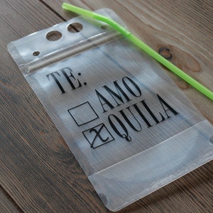 May include: A clear plastic pouch with a zip closure and a green straw sticking out of the top. The pouch has a black graphic that says "TE: AMO QUILLA" with a tick next to "QUILLA" and an empty box next to "AMO".