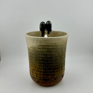 Puede incluir: Una taza de cerámica con un esmalte marrón y blanco. La taza tiene dos pequeñas asas en forma de pájaro negro en la parte superior.