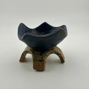 Puede incluir: Un pequeño cuenco de cerámica azul con un diseño único en forma de estrella se asienta sobre un soporte de tres patas hecho de cerámica marrón y beige moteada.