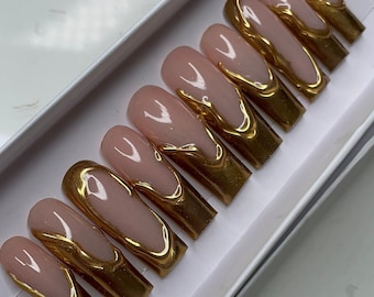 May include: A set of ten artificial nails in a white box. Each nail has a nude base with a gold metallic French tip design. The nails are long and rectangular, with a glossy finish.