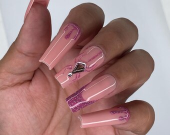 May include: A hand displaying long, square-shaped artificial nails. The nails are painted a light pink with glitter accents. Some nails feature a dripping glitter design, while others have a geometric design with a small gem.
