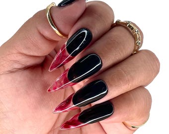 May include: A hand displaying long, black and red stiletto-shaped press-on nails. The nails have a glossy black base with a translucent red and white marbled design at the tips. Gold rings adorn the fingers.