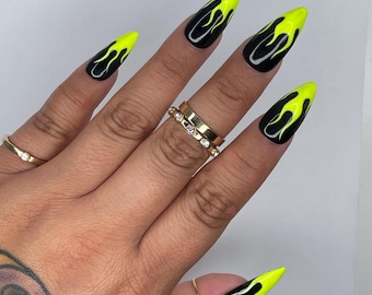 May include: Black stiletto nails with neon yellow flame designs. The nails are long and pointed, with a glossy finish. The hand also features gold rings and a tattoo on the arm.