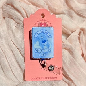 May include: A rectangular badge reel with a blue and white design featuring a cartoon bear holding a heart, with the text "TODAY IS MY GRUMPY DAY!". The badge reel is attached to a pink card with the text "COCO'S CRAFTROOM".