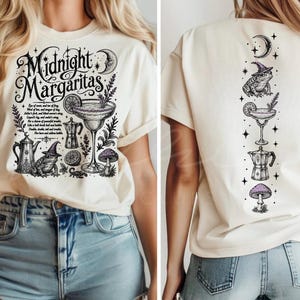 May include: A cream-colored t-shirt featuring black ink illustrations. The front displays the text Midnight Margaritas with a margarita glass, a toad in a witch hat, a coffee pot, and a mushroom. The back shows a vertical arrangement of these icons.